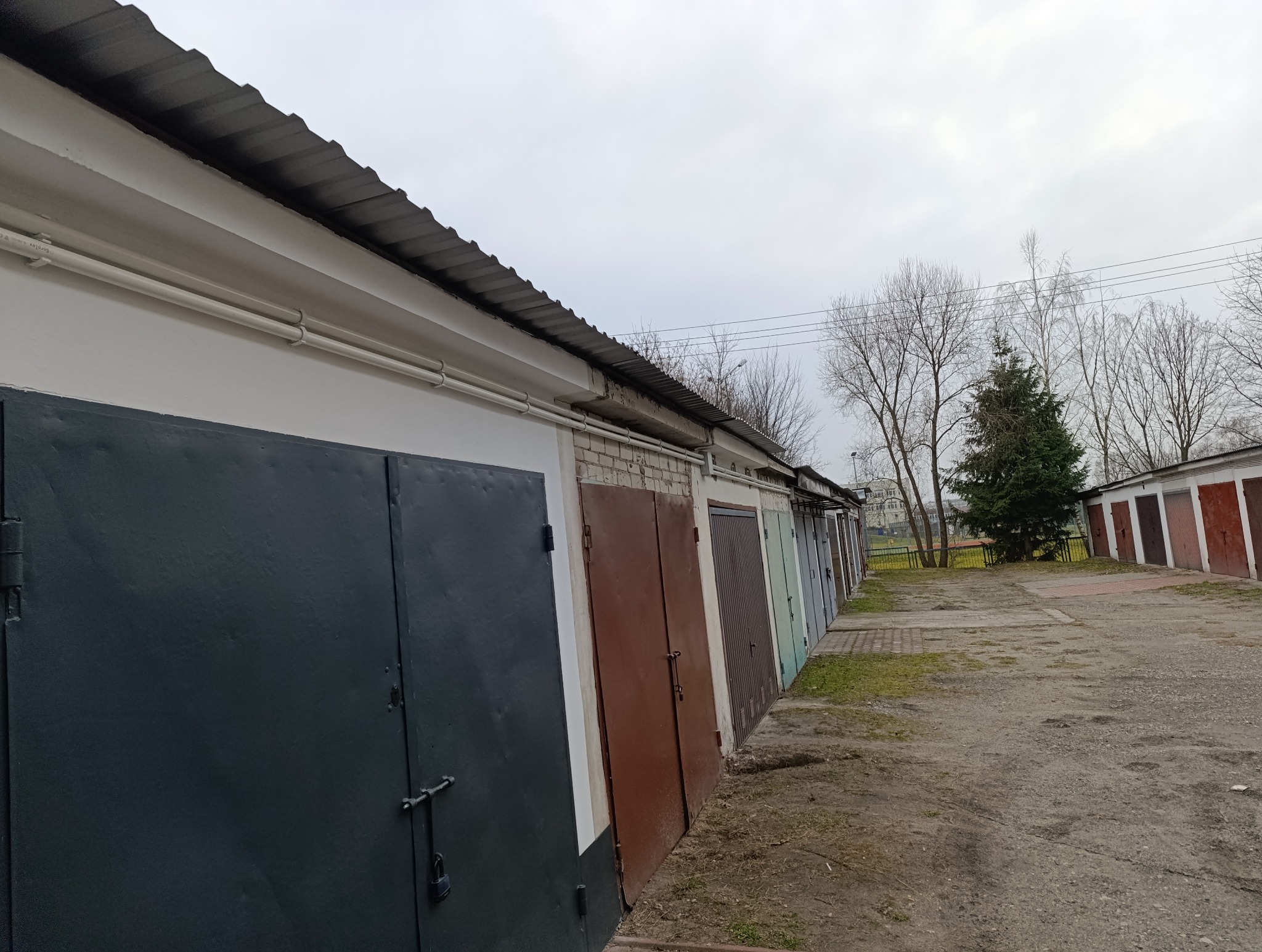 Rząd garaży z metalowymi drzwiami w różnych kolorach, widoczne rury instalacji elektrycznej pod dachem, pochmurne niebo w tle.