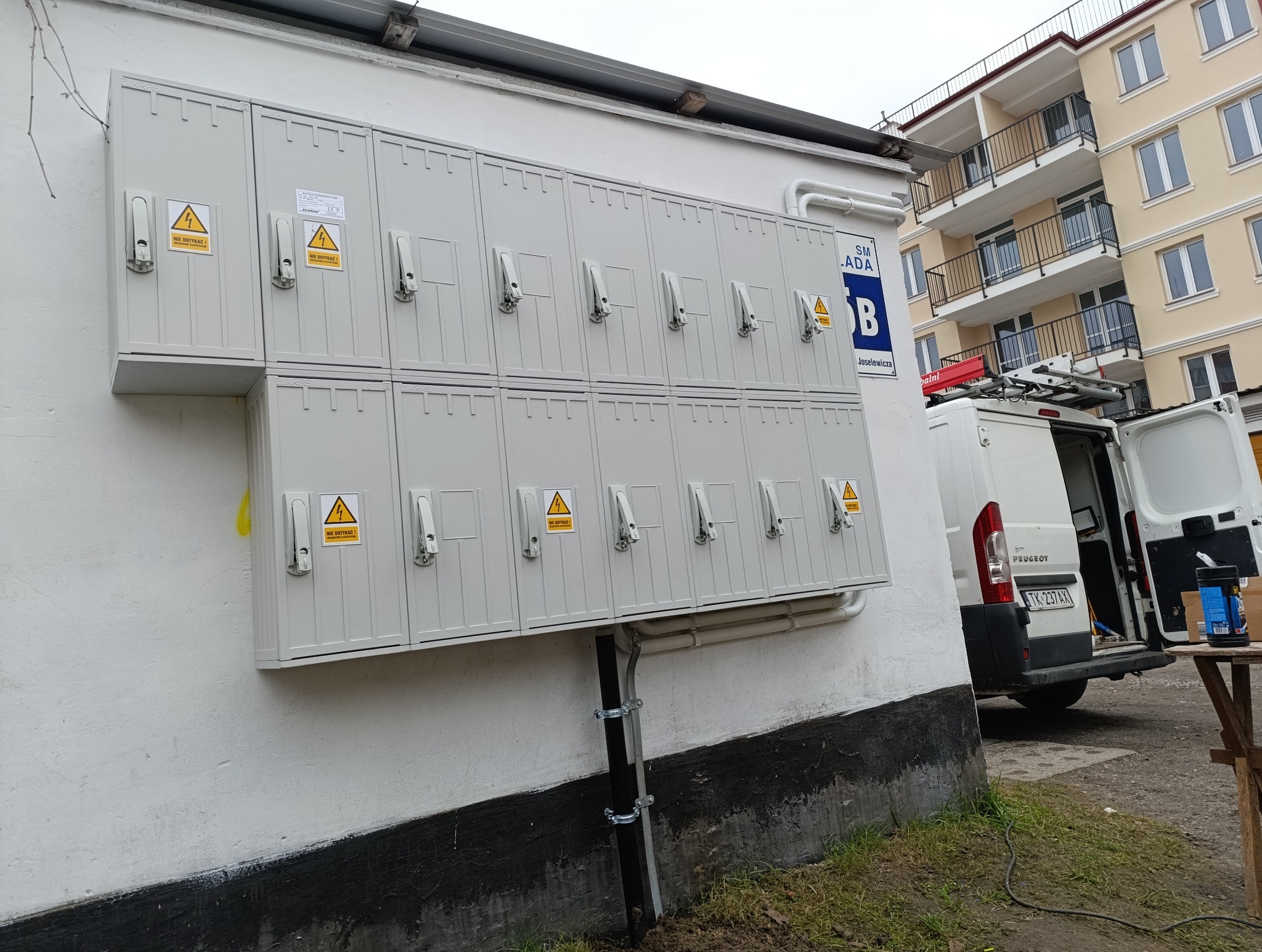 Zestaw połączonych szarych skrzynek elektrycznych z ostrzeżeniami 'Nie dotykać!' na ścianie budynku, z zaparkowanym białym vanem serwisowym w tle i fragmentem nowego budynku mieszkalnego