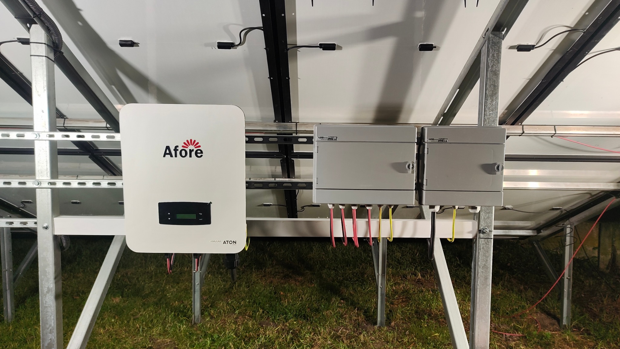 Widok od dołu na zamontowaną instalację fotowoltaiczną z widocznym inwerterem marki Afore i skrzynkami przyłączeniowymi, ustawioną na metalowej konstrukcji na tle trawy.