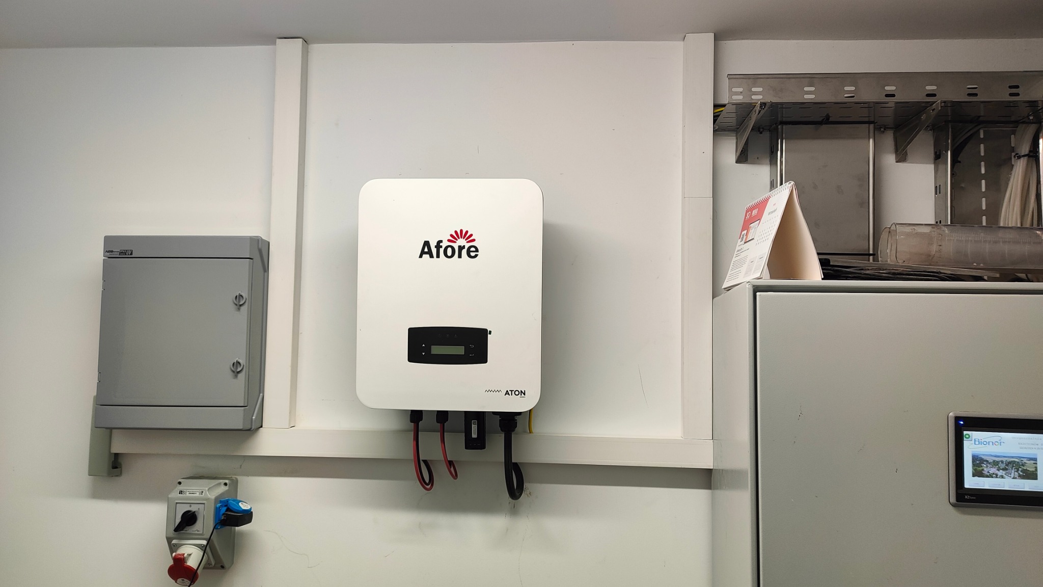 Biały falownik Afore zamontowany na ścianie w pomieszczeniu technicznym, obok szara skrzynka elektryczna i gniazdo siłowe. Widoczne kable zasilające. W tle metalowe korytka kablowe.