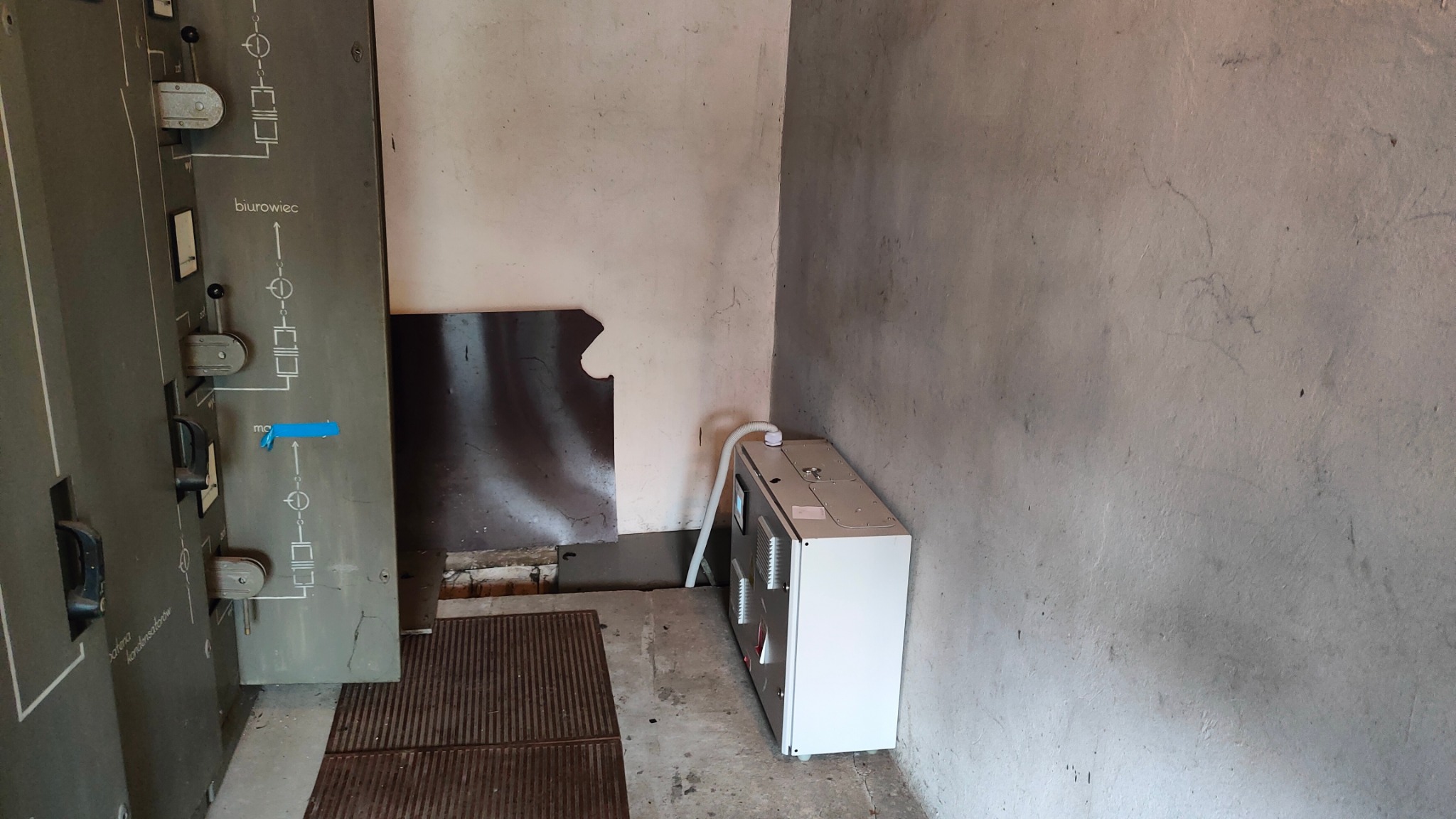 Wnętrze pomieszczenia z szarą, betonową ścianą, w którym znajduje się stara szafa elektryczna z opisem 'biurowiec' i nowa, biała jednostka klimatyzacyjna z podłączonym przewodem. Widoczna podłoga...