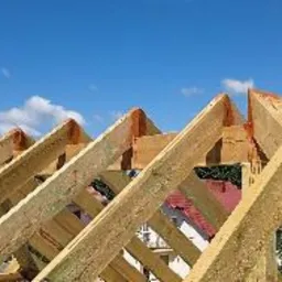 Konstrukcja drewniana dachu w trakcie budowy, widoczne elementy więźby dachowej na tle błękitnego nieba.