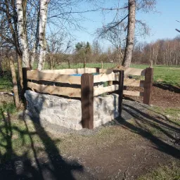Obrzeże studni z kamienia i drewna w ogrodzie, widoczne brzozy i trawnik w tle.