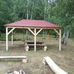 Drewniana altana z czerwonym dachem w lesie brzozowym, z surowym stołem i ławkami z bali, teren lekko nierówny.