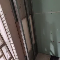 Montaż konstrukcji z płyt karton-gips w Poznaniu: widoczne metalowe profile, częściowo zamontowane płyty w kolorze zielonym, oraz fragment rusztowania. Otwór w podłodze sugeruje prace instalacyjne...