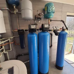 Instalacja uzdatniania wody z trzema niebieskimi zbiornikami ciśnieniowymi i osprzętem hydraulicznym w pomieszczeniu technicznym w Baranowie Sandomierskim, widoczne również zbiorniki wyrównawcze...