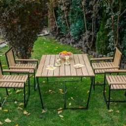 Zestaw mebli ogrodowych: kwadratowy stół i cztery krzesła z drewnianymi siedziskami i oparciami na czarnych metalowych ramach, ustawione na trawniku z opadłymi liśćmi, z owocami i filiżankami...