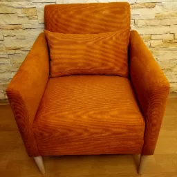 Pomarańczowy fotel z poduszką, obity tkaniną w prążki, na tle ściany z kamienia dekoracyjnego.