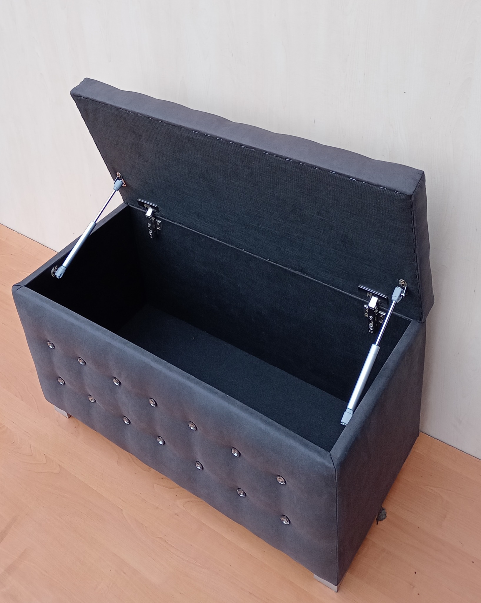 Otwierana, tapicerowana skrzynia w kolorze grafitowym z dekoracyjnymi guzikami, podparta na metalowych nóżkach, z widocznymi zawiasami i siłownikami.