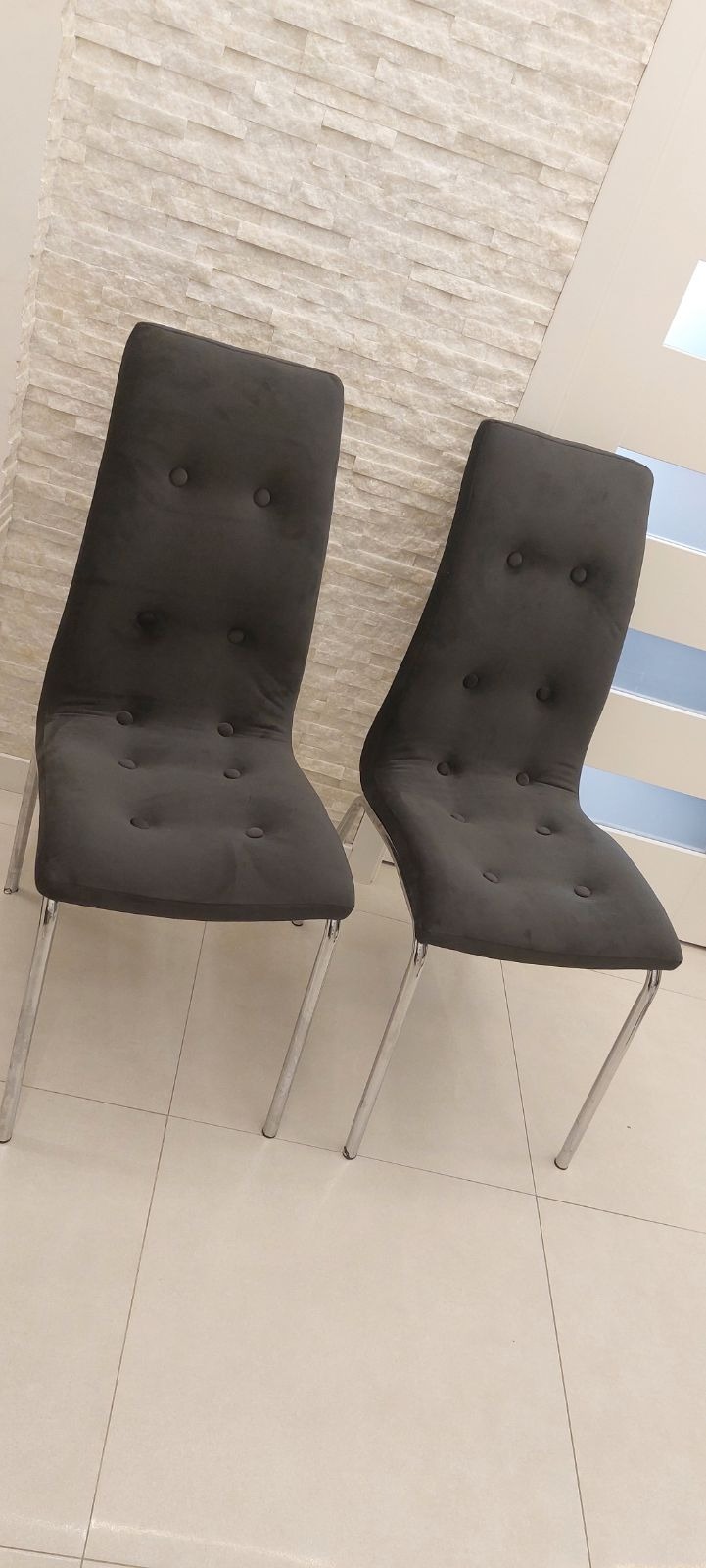 Dwa krzesła tapicerowane ciemnoszarą tkaniną z guzikami, na chromowanych nogach, ustawione na jasnej podłodze z płytek, na tle ściany z dekoracyjnej cegły.