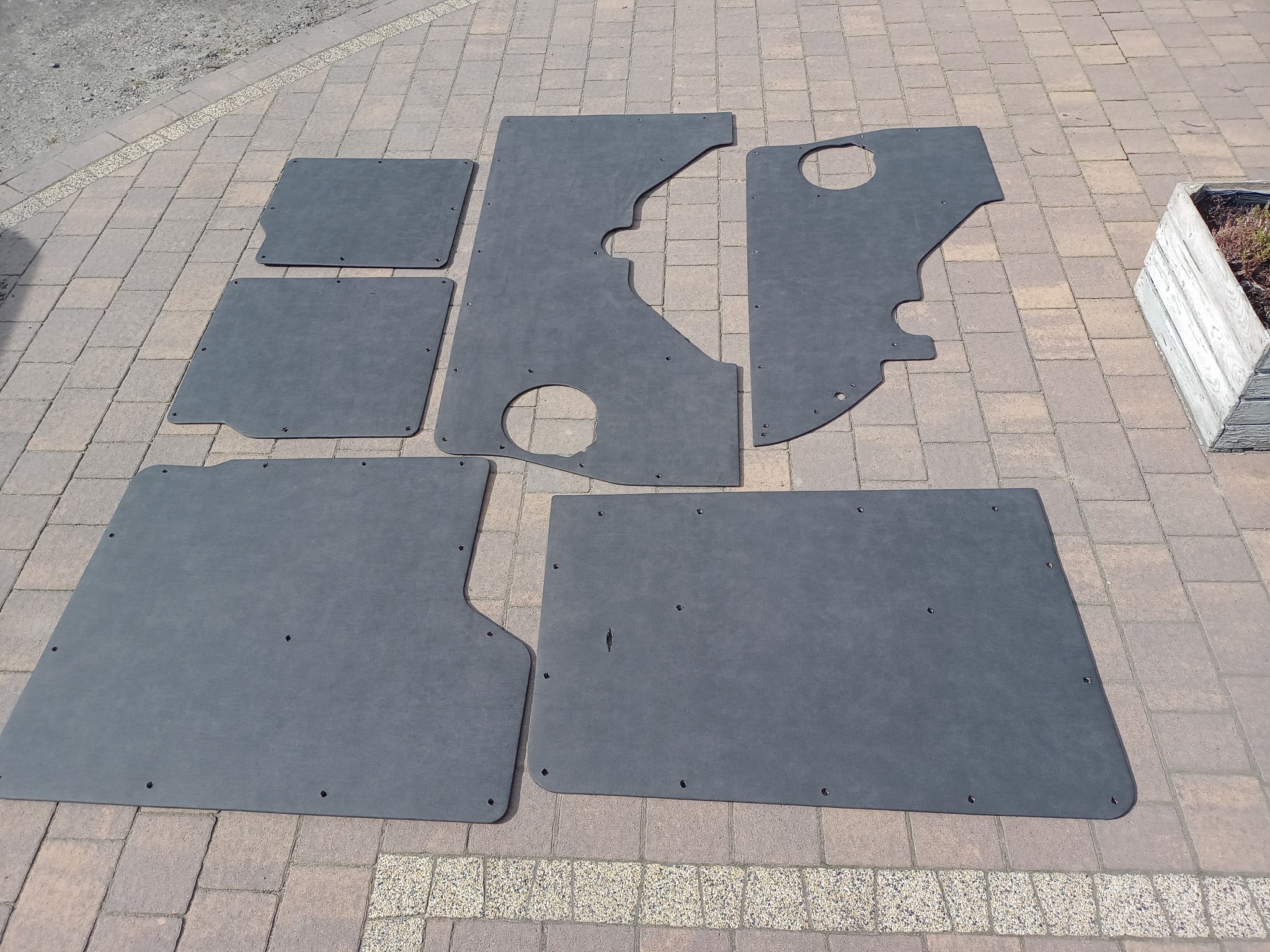 Elementy tapicerki samochodowej z otworami montażowymi, rozłożone na brukowanej powierzchni.