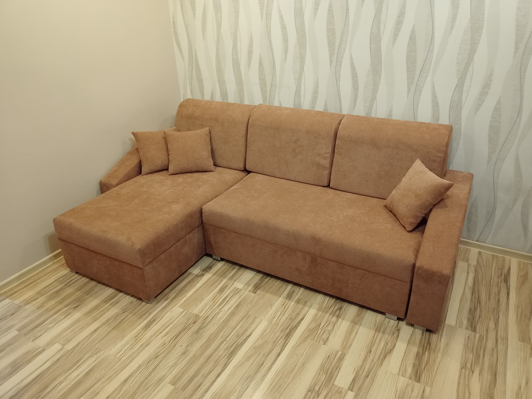 Rozłożona, narożna kanapa w odcieniach brązu z dwiema poduszkami na siedzisku i dwiema na szezlongu, na drewnianej podłodze, na tle ściany w biało-szare wzory.