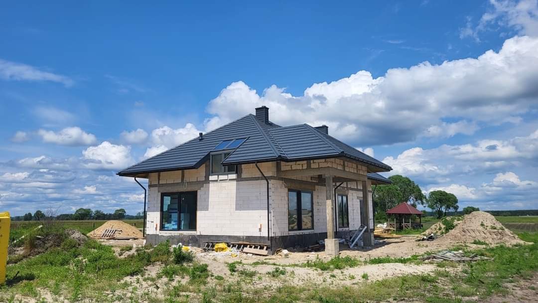 Dom jednorodzinny w trakcie budowy, z szarą, skośną konstrukcją dachu z oknem dachowym, widoczne pustaki, nieotynkowane ściany, w tle błękitne niebo z chmurami i zielony krajobraz. Na dachu...