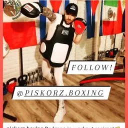 Mężczyzna w czapce i ochraniaczu na korpusie, trzymający tarcze bokserskie w rękach, w tle flagi Meksyku i Polski, oraz sprzęt do ćwiczeń.