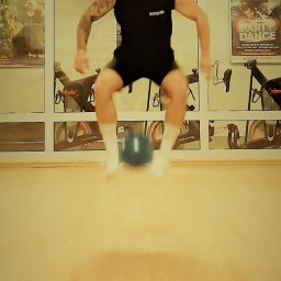 Warmiński Klub Bokserski Piskorz - Mężczyzna w czarnym stroju i czerwonej bandanie skacze nad niebieskim kettlebellem w siłowni z rowerami stacjonarnymi w tle.