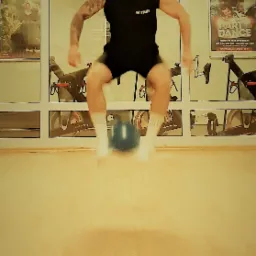 Mężczyzna w czarnym stroju i czerwonej bandanie skacze nad niebieskim kettlebellem w siłowni z rowerami stacjonarnymi w tle.