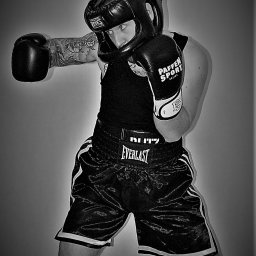 Warmiński Klub Bokserski Piskorz - Czarno-białe zdjęcie boksera w pełnym stroju ochronnym, z tatuażami na ramieniu, w pozycji gotowej do walki na tle szarej ściany.