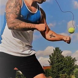 Warmiński Klub Bokserski Piskorz - Mężczyzna z bandaną na głowie, trenuje refleks uderzając w piłkę tenisową przymocowaną do elastycznej linki na tle nieba i zieleni