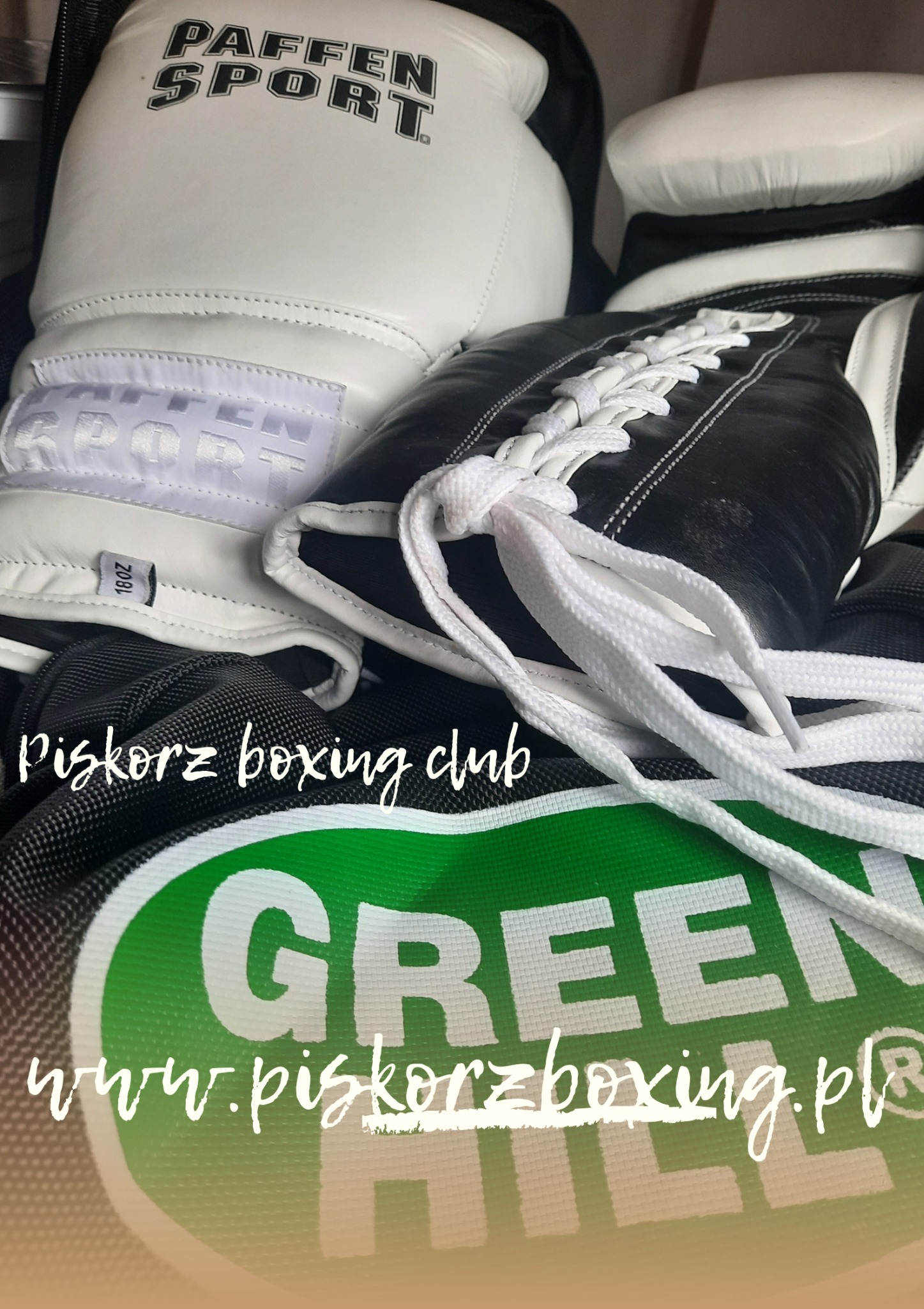 Para rękawic bokserskich Paffen Sport, biało-czarnych, z białymi sznurówkami, leżąca na torbie sportowej Green Hill z logo Piskorz Boxing Club i adresem strony internetowej.