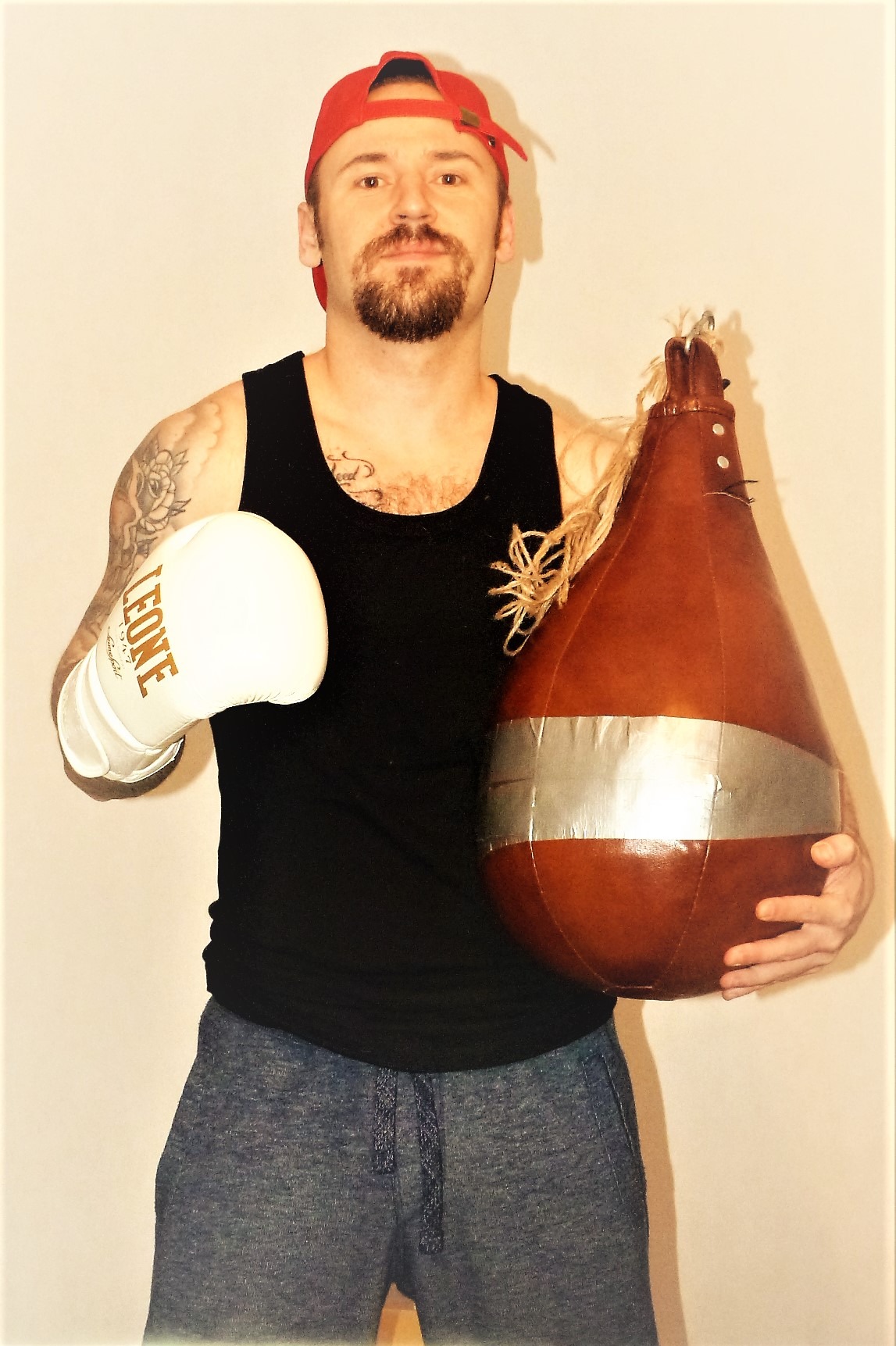 Mężczyzna z brodą w czerwonej czapce trzyma skórzaną gruszkę bokserską z taśmą i białą rękawicę bokserską Leone na tle jasnej ściany.