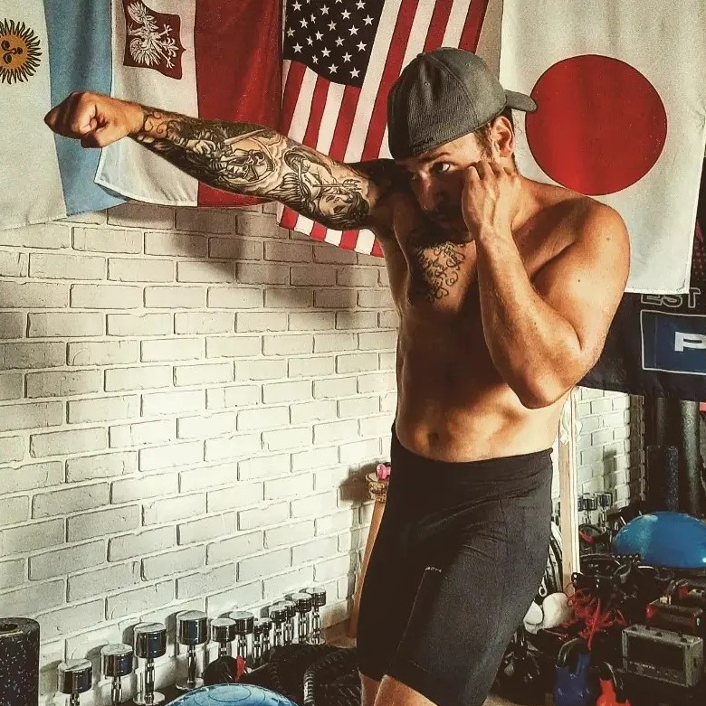 Mężczyzna w czapce i spodenkach, z tatuażami na ramieniu, w pozycji bokserskiej na tle ściany z cegły i flag Argentyny, Polski, USA i Japonii.