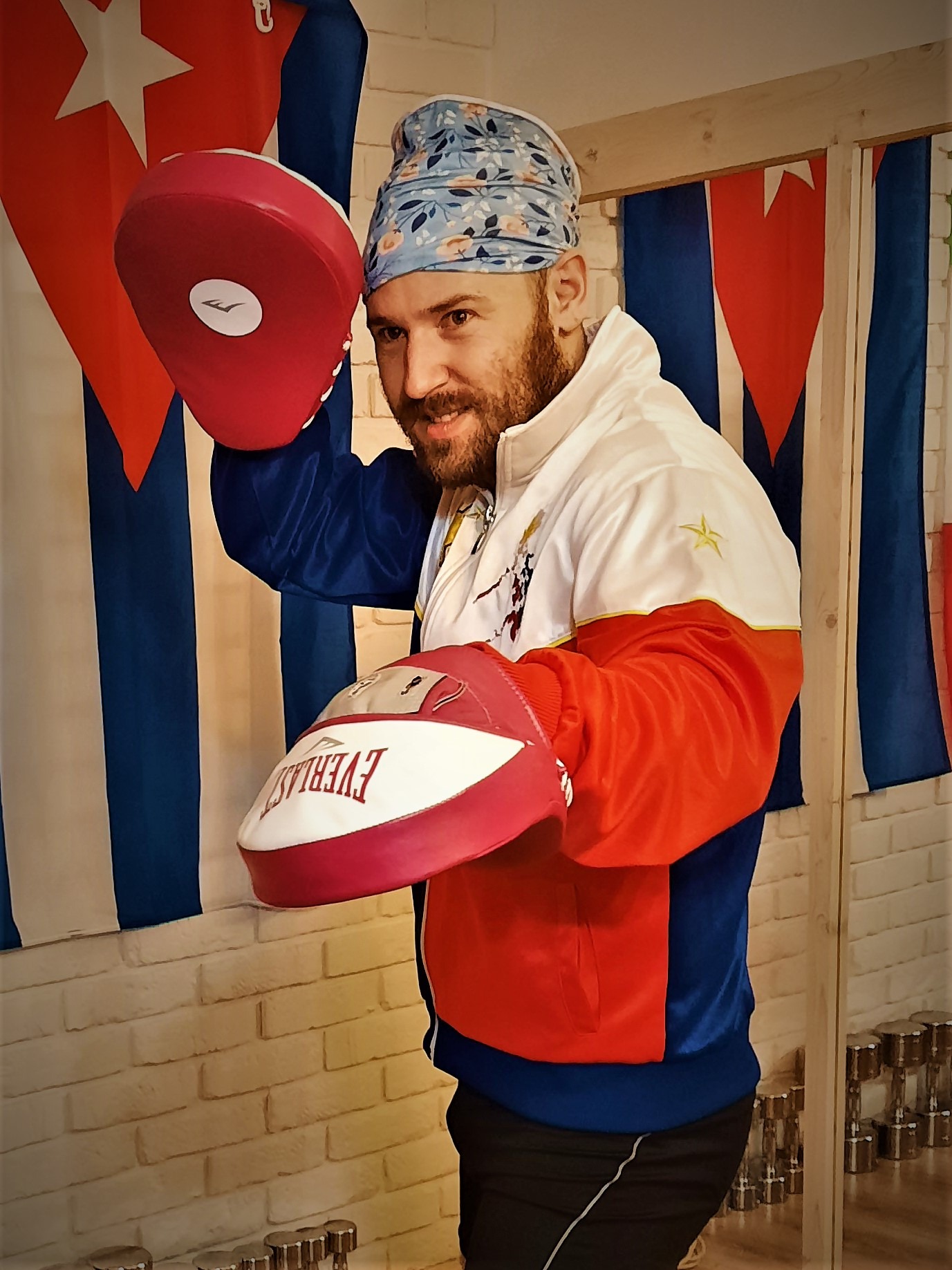 Uśmiechnięty mężczyzna z brodą w sportowej kurtce i wzorzystej bandanie, trzymający czerwone tarcze bokserskie Everlast na tle flagi Kuby i białej ceglanej ściany, w tle widoczne hantle.