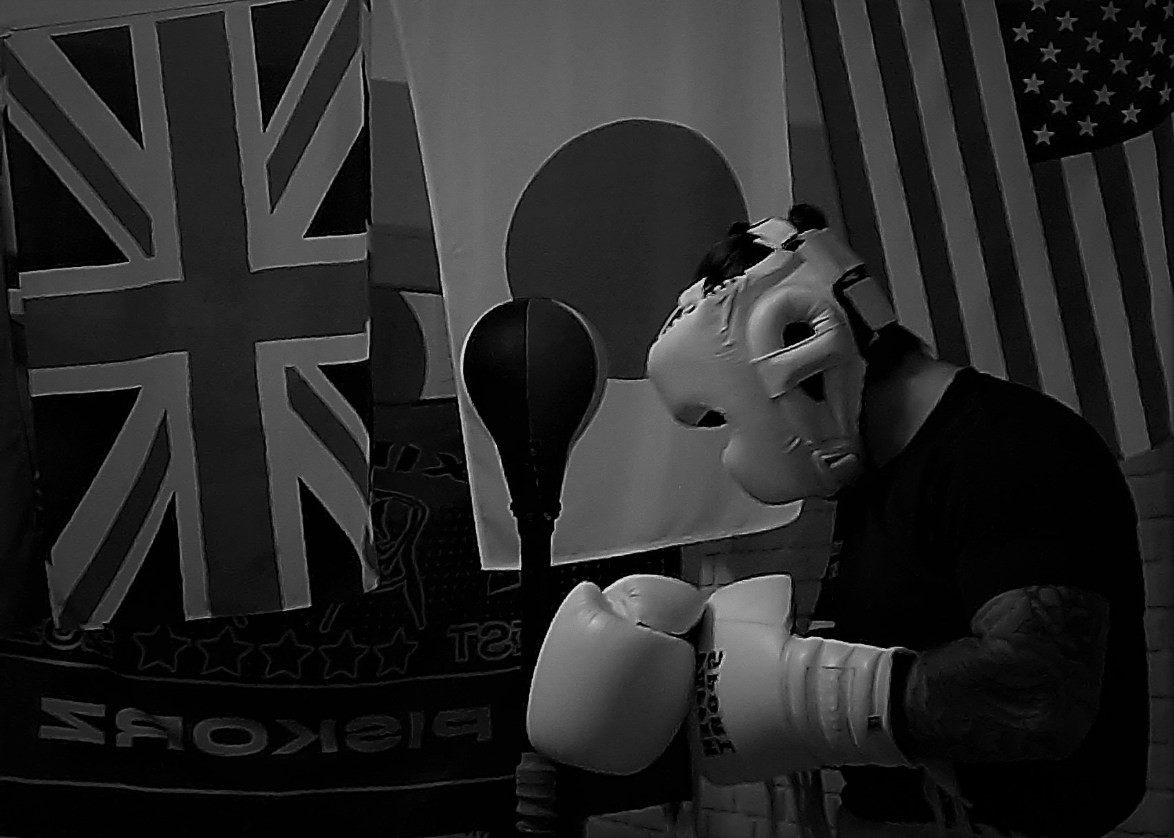 Czarno-białe zdjęcie boksera w kasku i rękawicach, z flagami Wielkiej Brytanii, Japonii i USA w tle oraz gruszką bokserską