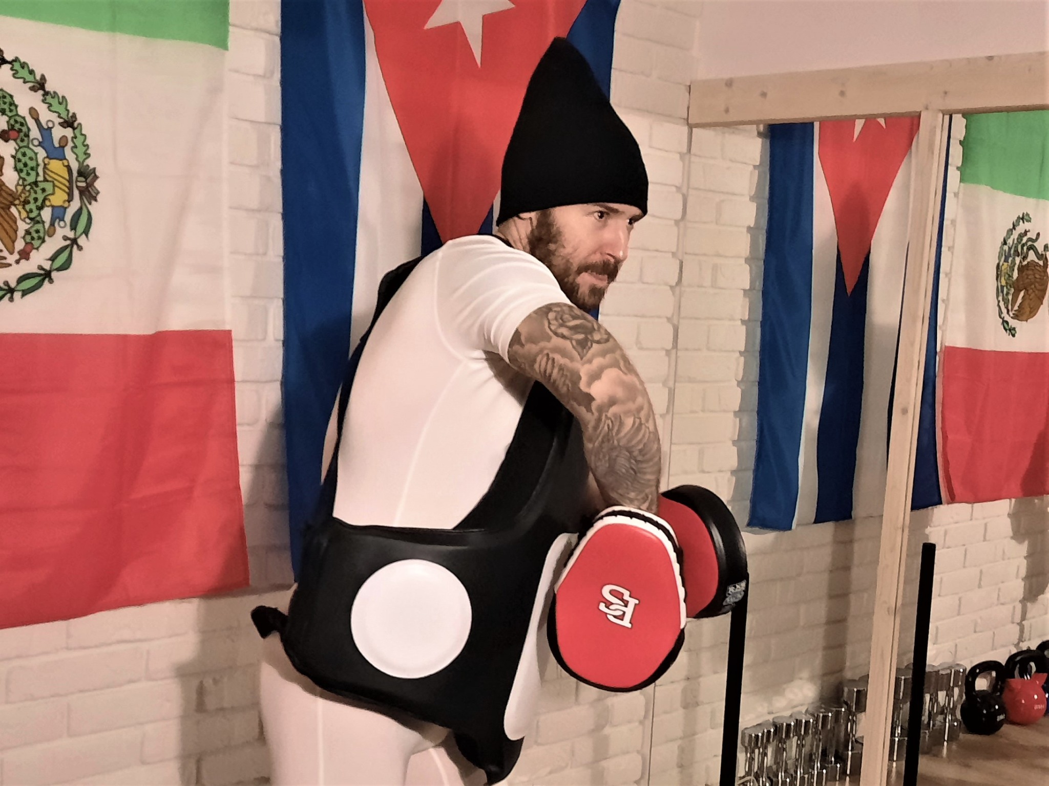 Mężczyzna z brodą i tatuażem na ramieniu w czapce, białej koszulce i ochraniaczu na korpusie, trzymający czerwone łapy trenerskie na tle flag Kuby i Meksyku, podczas treningu sportów walki.