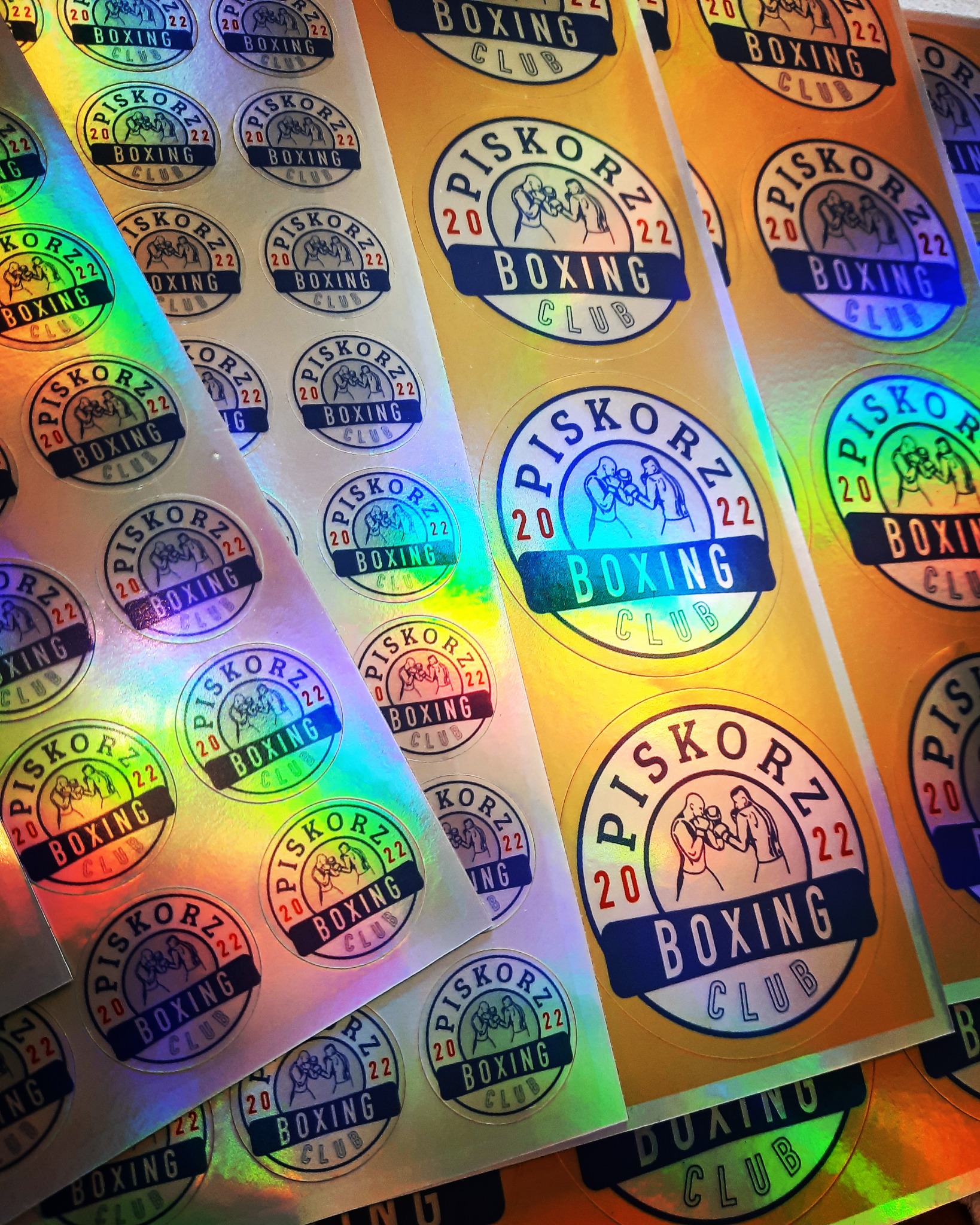 Arkusz naklejek z okrągłym logo Piskorz Boxing Club z grafiką dwóch bokserów w ringu, datą 2022 i nazwą klubu, widoczne efekty holograficzne i złote tło.