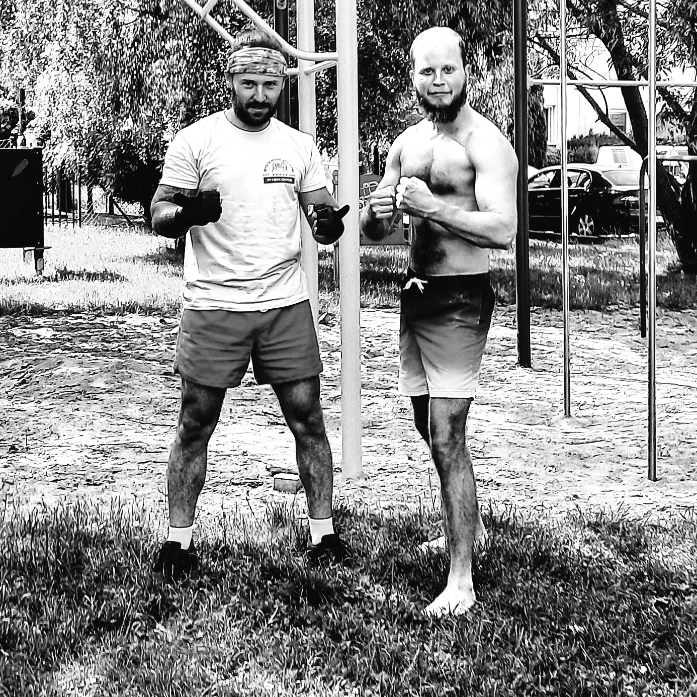 Dwóch mężczyzn w sportowych strojach pozuje na tle plenerowej siłowni, jeden w rękawicach bokserskich i opasce na głowie, drugi z zaciśniętymi pięściami i gołą klatką piersiową.