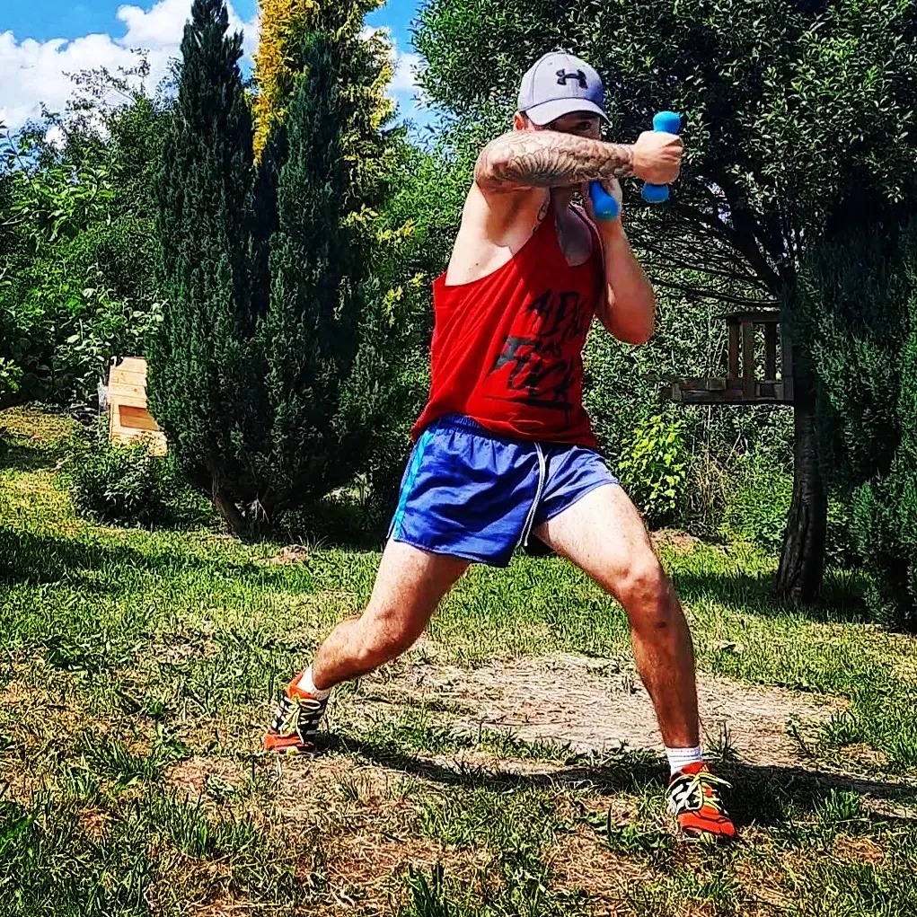 Mężczyzna w czerwonym tank topie i niebieskich spodenkach ćwiczy boks z małymi niebieskimi hantlami na trawniku przed drzewami.