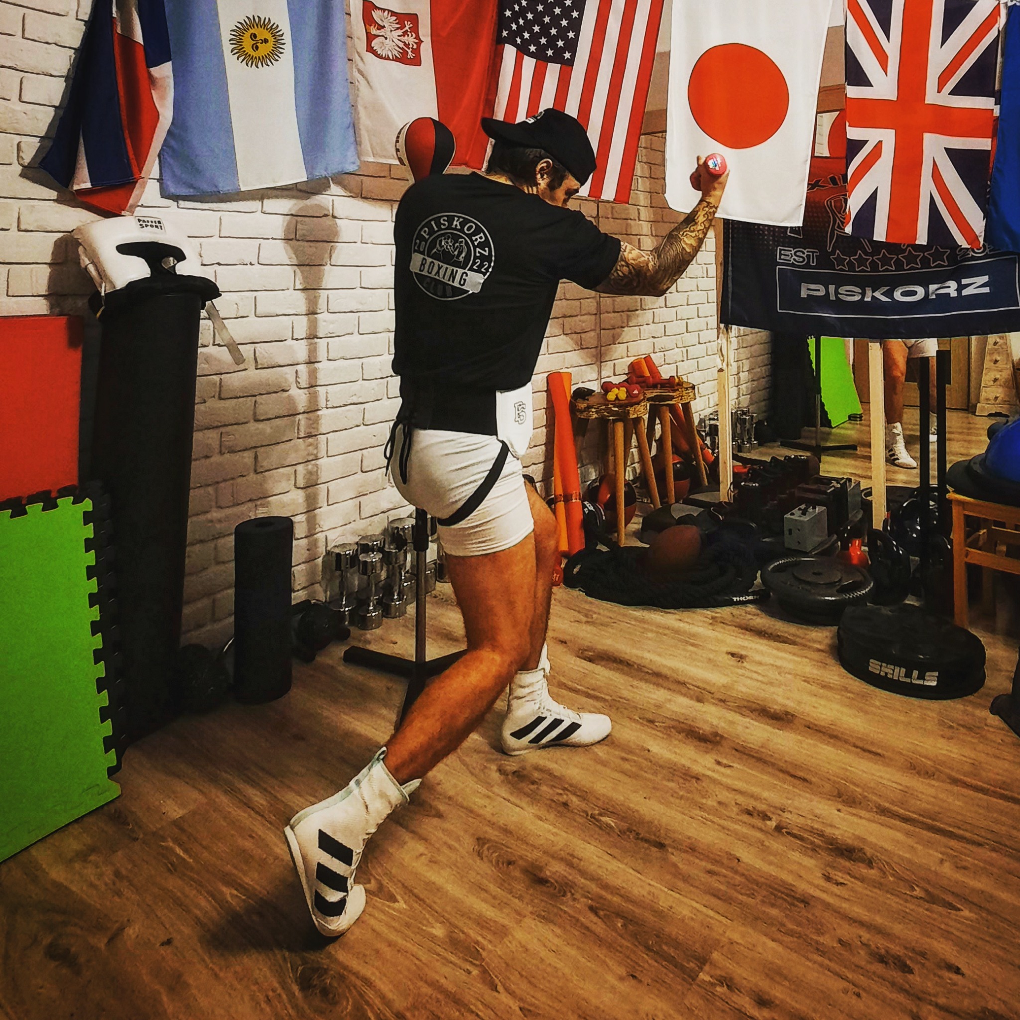 Mężczyzna w stroju bokserskim, z tatuażami na ramionach, trenuje w domowej siłowni z flagami różnych krajów na ścianie, wykonując uderzenie z małą piłeczką.