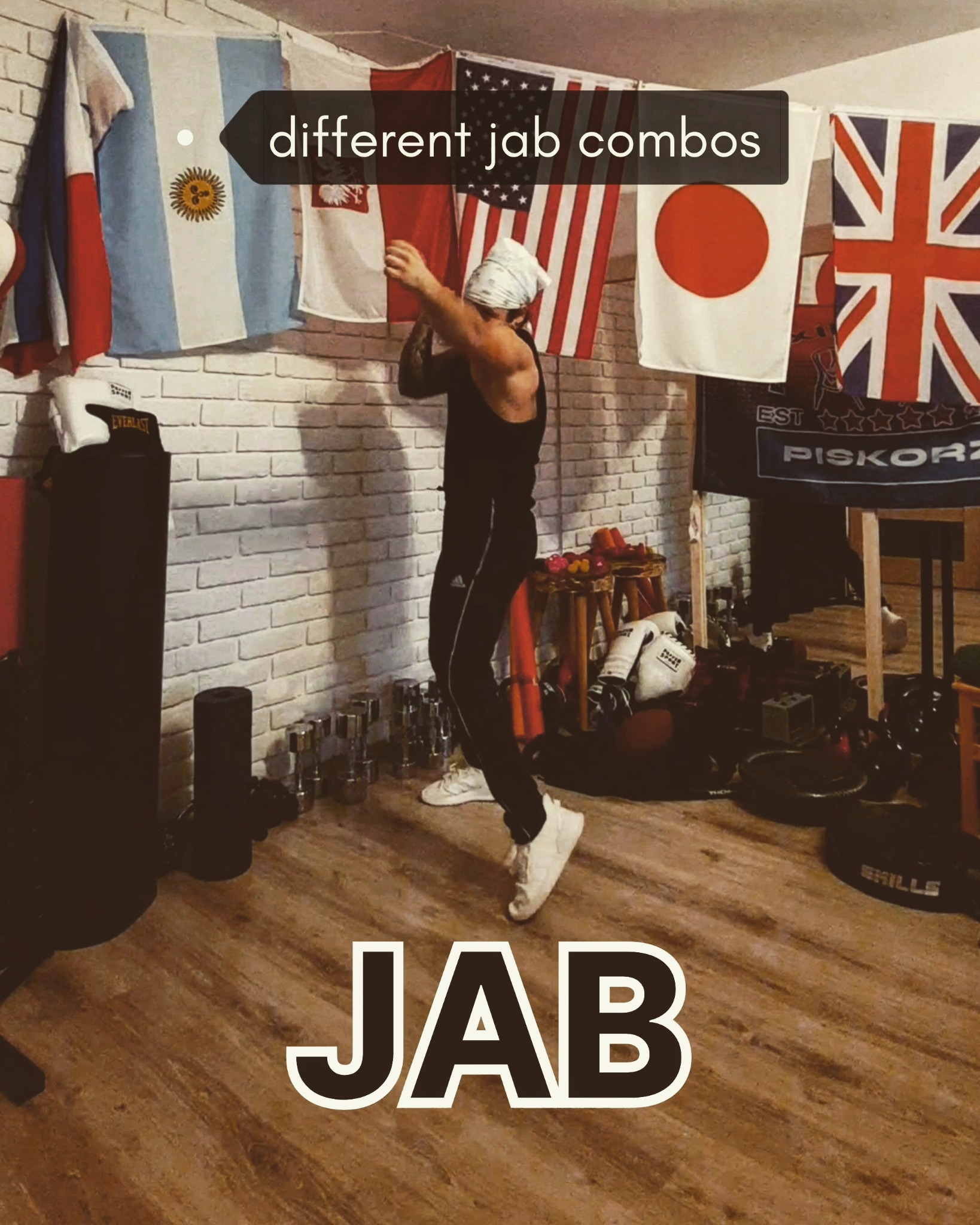 Mężczyzna w czarnym stroju i bandamie na głowie wykonuje uderzenie pięścią (jab) na tle ściany z flagami różnych państw, w tle widoczne worki treningowe, hantle i inny sprzęt sportowy.