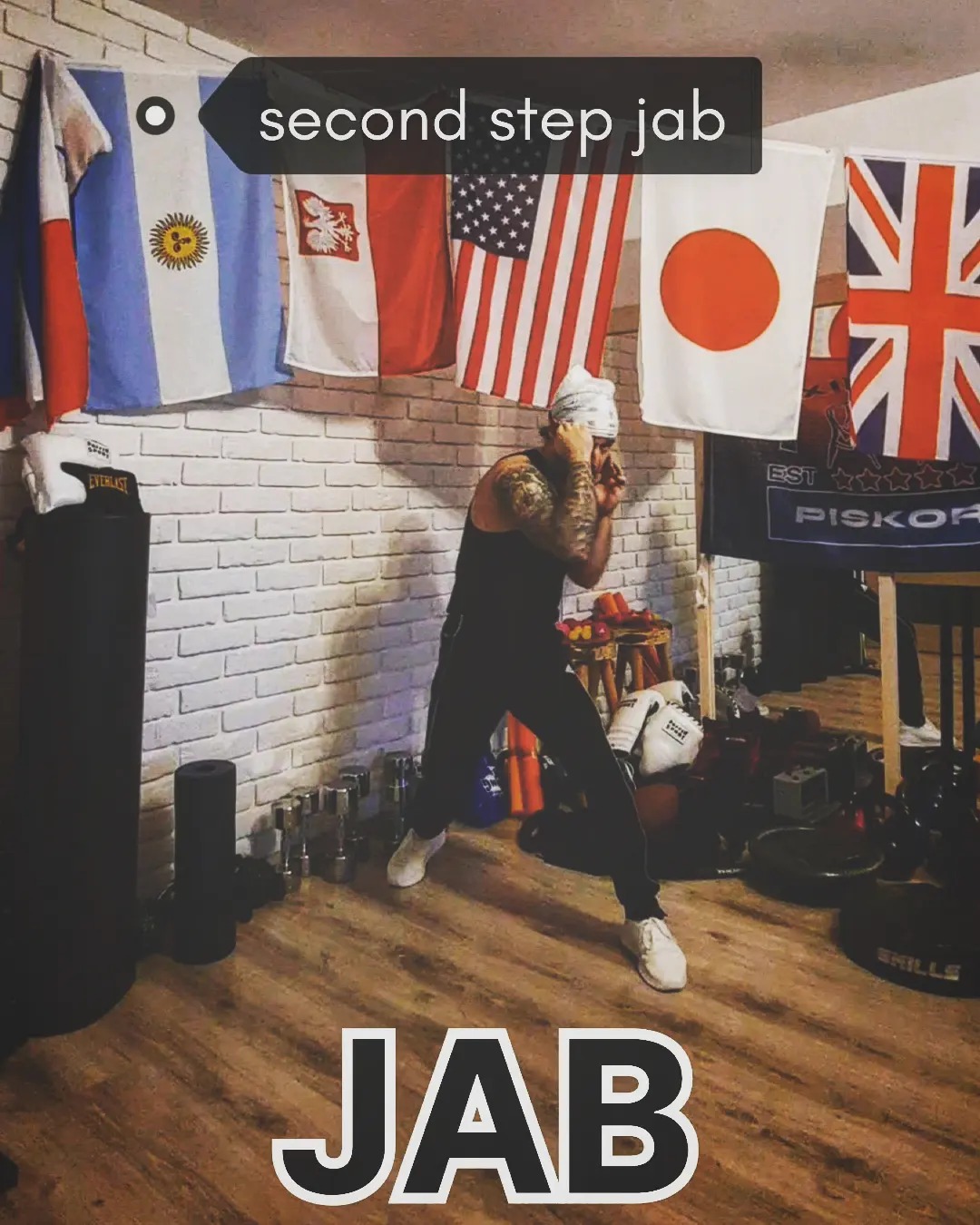 Mężczyzna z tatuażami w stroju sportowym wykonuje cios bokserski (jab) w domowej siłowni, na ścianie wiszą flagi różnych państw.