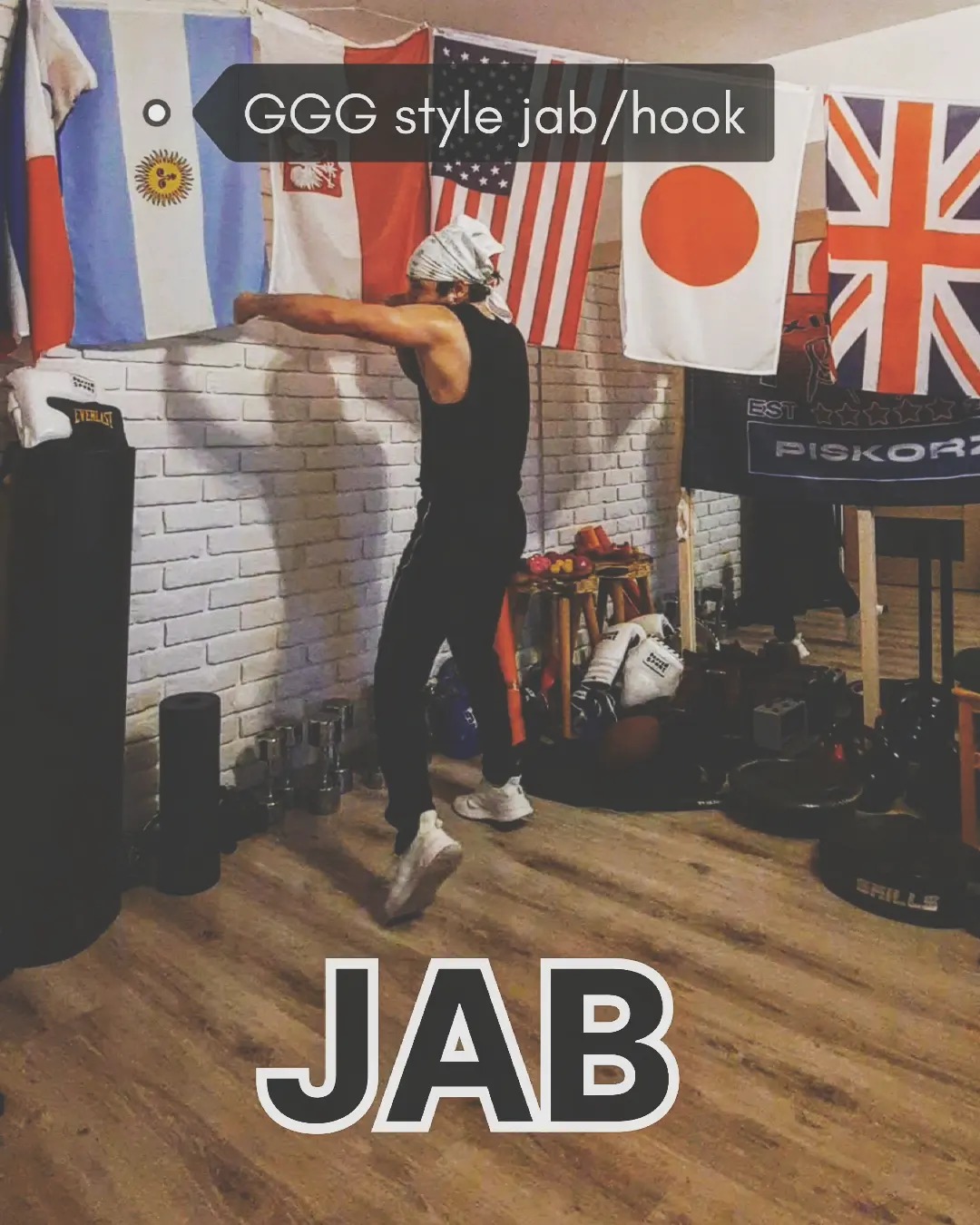 Mężczyzna w czarnym stroju treningowym wykonuje jab w sali bokserskiej z flagami różnych krajów w tle.