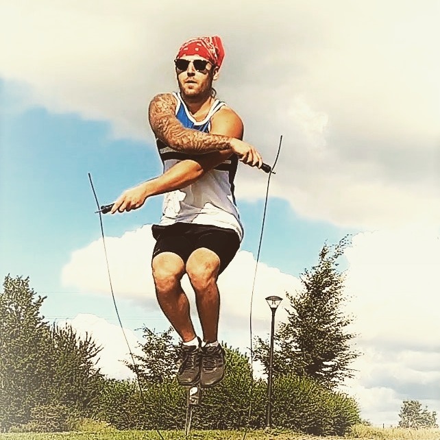 Mężczyzna w bandanie i okularach przeciwsłonecznych skacze na skakance na tle błękitnego nieba z chmurami i zieleni