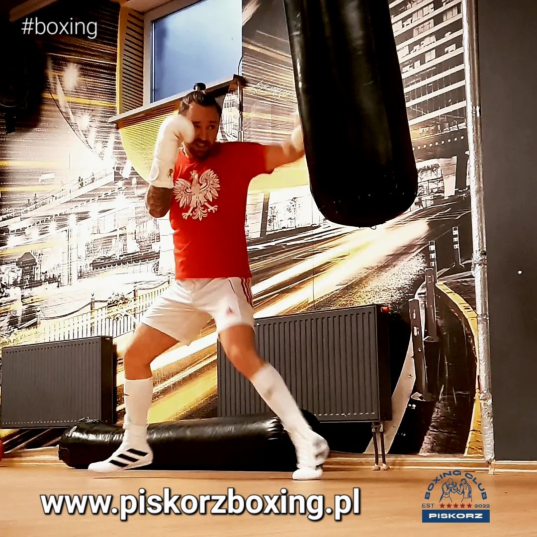 Mężczyzna w czerwonej koszulce z orłem, białych spodenkach i rękawicach bokserskich uderza w czarny worek treningowy w sali bokserskiej z grafiką miejską na ścianie.
