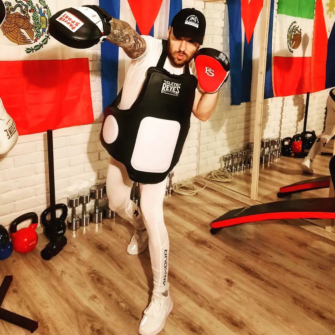 Mężczyzna w białym stroju treningowym, ochraniaczu na korpus i rękawicach bokserskich pozuje w pomieszczeniu z flagami, hantlami i kettlami.