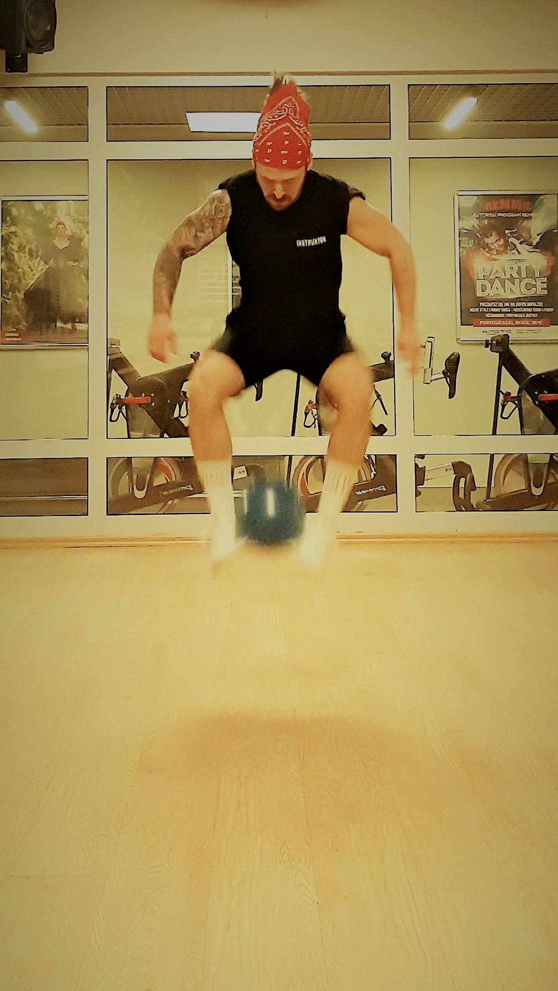 Mężczyzna w czarnym stroju i czerwonej bandanie skacze nad niebieskim kettlebellem w siłowni z rowerami stacjonarnymi w tle.