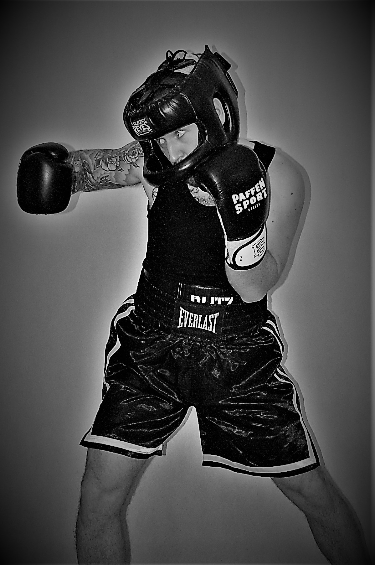 Czarno-białe zdjęcie boksera w pełnym stroju ochronnym, z tatuażami na ramieniu, w pozycji gotowej do walki na tle szarej ściany.