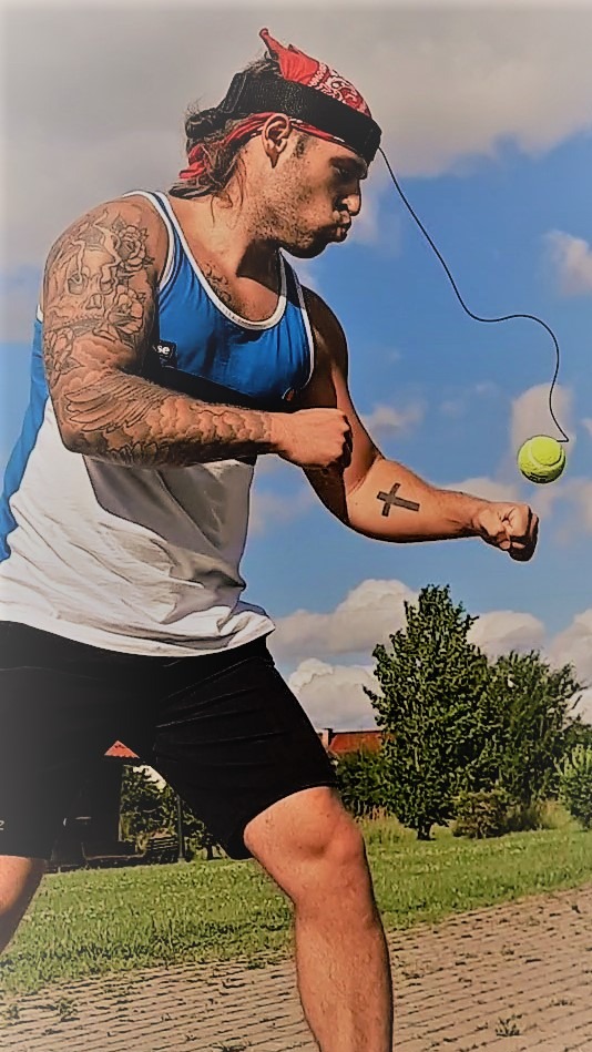 Mężczyzna z bandaną na głowie, trenuje refleks uderzając w piłkę tenisową przymocowaną do elastycznej linki na tle nieba i zieleni