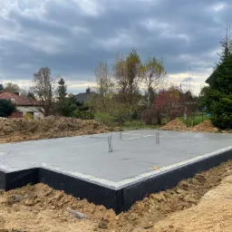 Wylana płyta fundamentowa z hydroizolacją pionową, wystające pręty zbrojeniowe, przygotowana pod budowę domu jednorodzinnego, widok na działkę budowlaną w pochmurny dzień.