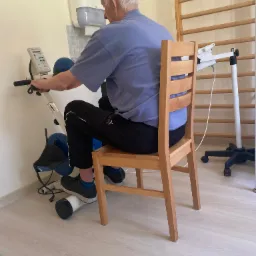 Pacjent w Jelcz-Laskowicach ćwiczy na rowerku rehabilitacyjnym, siedząc na drewnianym krześle; drabinki gimnastyczne w tle.