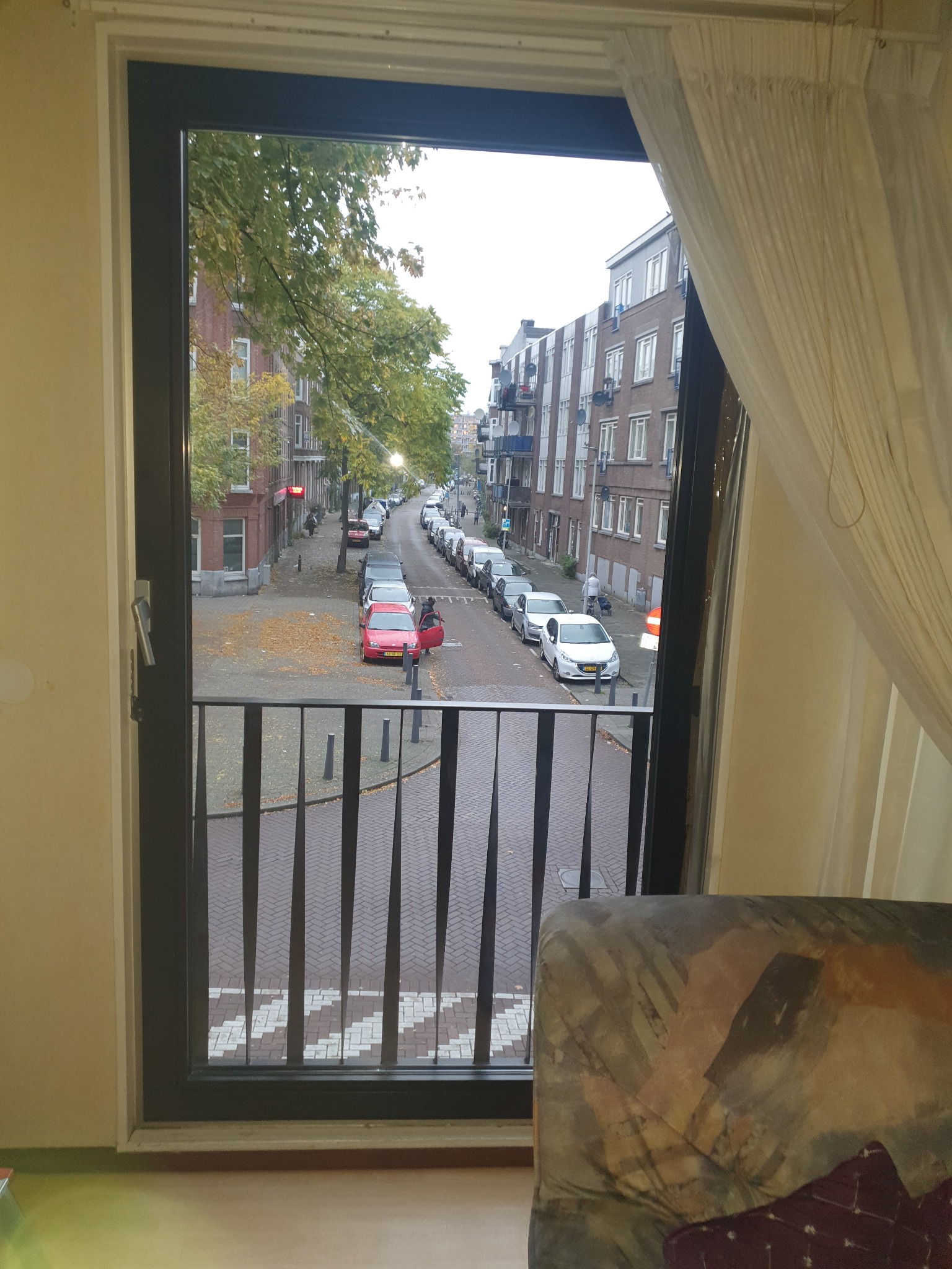 Widok z okna balkonowego na ulicę z zaparkowanymi samochodami, widoczne jesienne liście na chodniku i zasłona po prawej stronie.