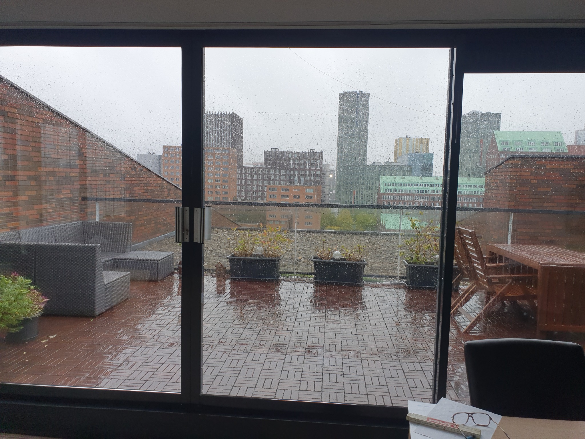 Widok z okna na mokry taras z meblami ogrodowymi, donicami z roślinami i panoramą miasta w tle w deszczowy dzień. Krople deszczu na szybie okna.