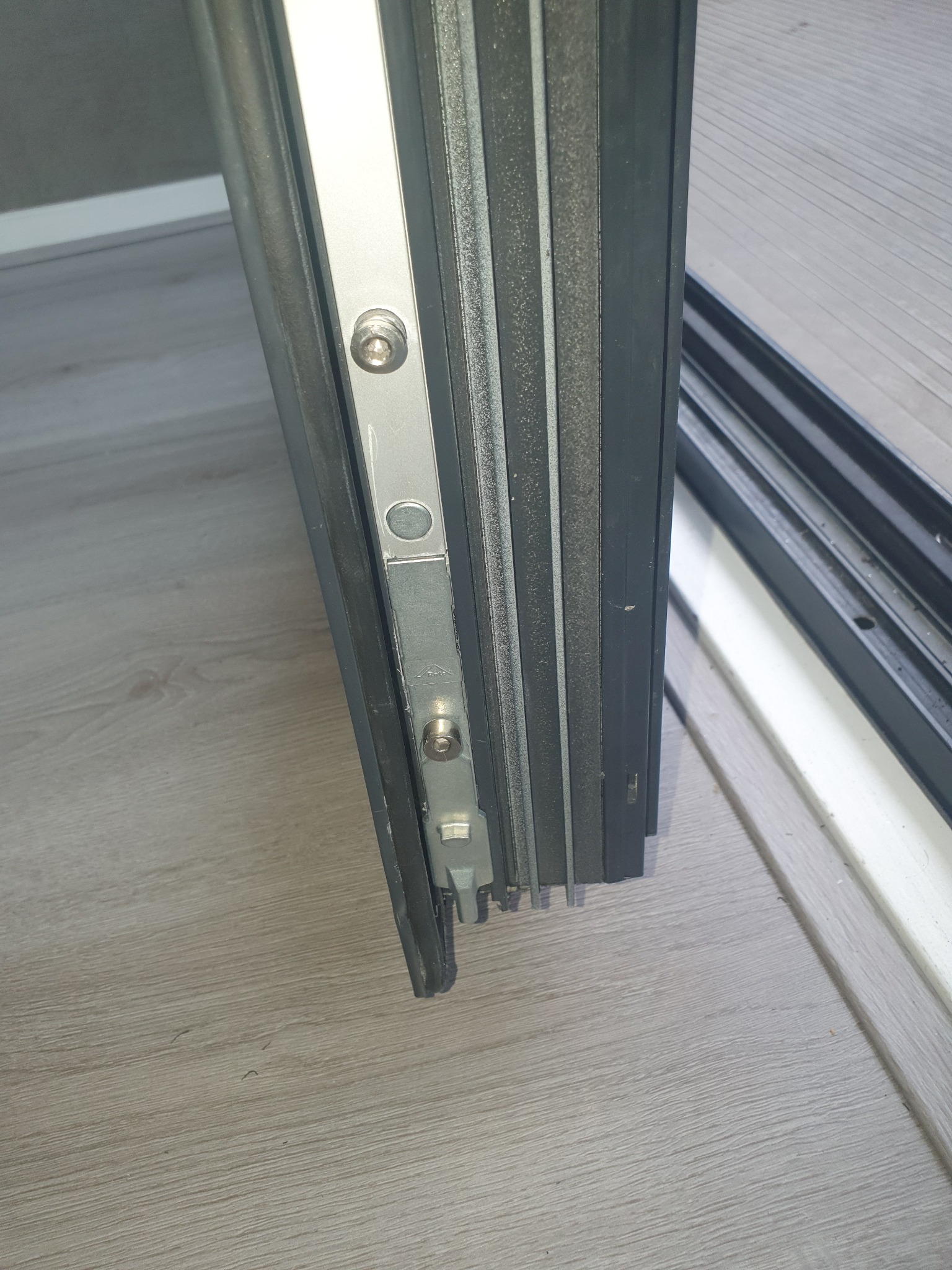 Detal metalowego okucia z dwoma śrubami w ramie okiennej, widoczne uszczelki i fragment podłogi z paneli.