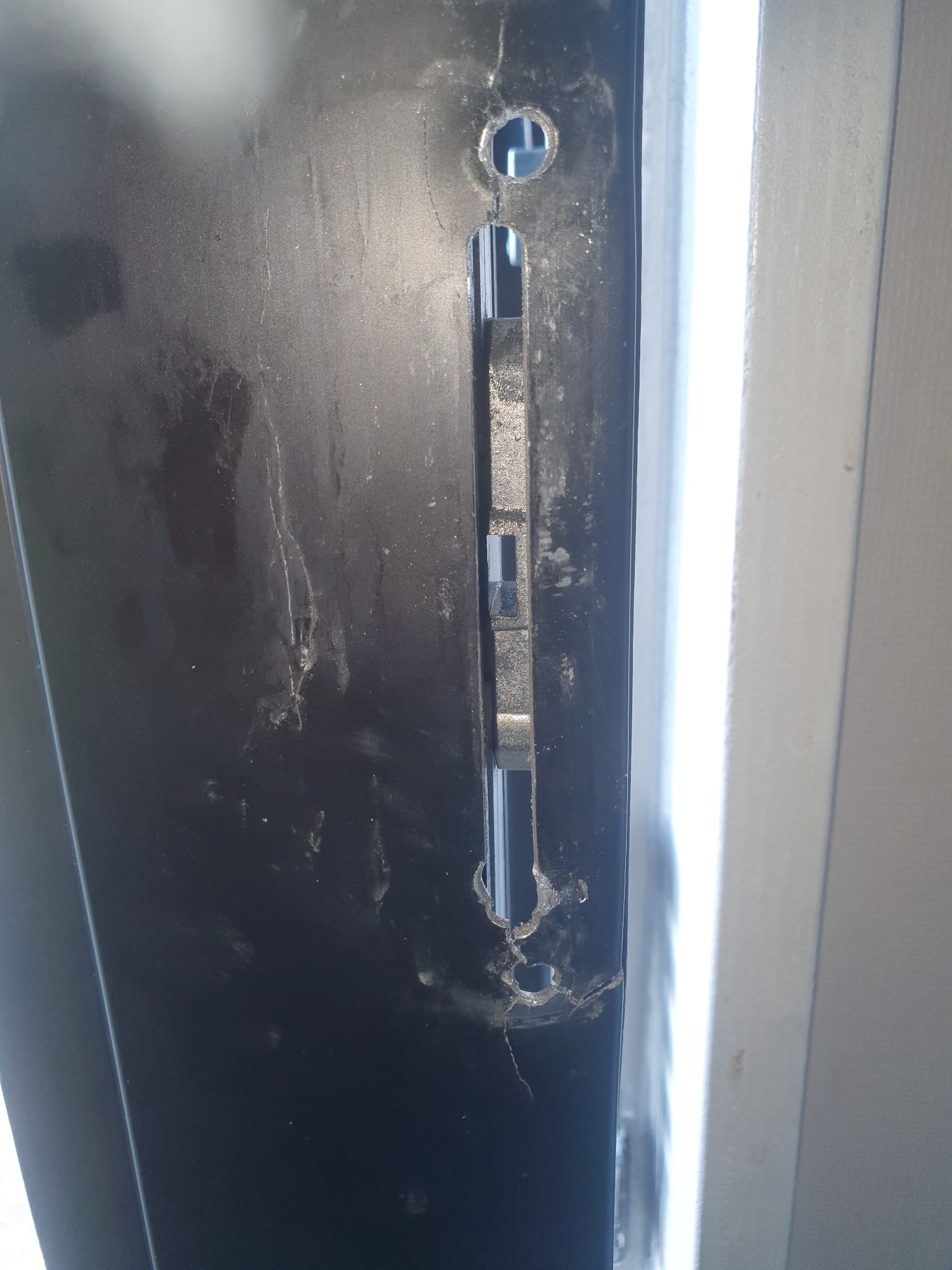 Czarny profil okienny z wywierconymi otworami i widocznym mechanizmem zamka, ślady uszkodzeń wokół otworu.