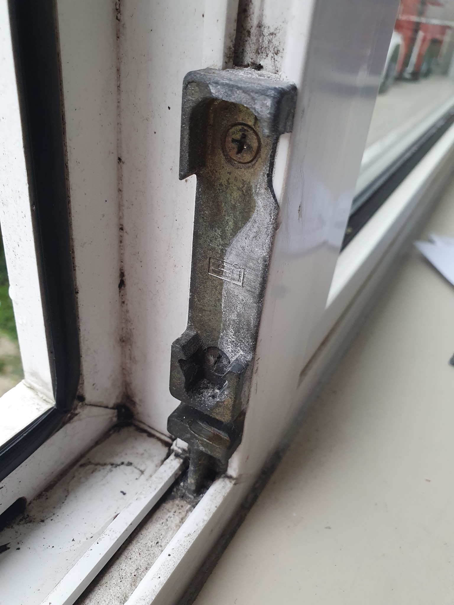 Stare, brudne okno PCV z widocznym, skorodowanym metalowym elementem mocującym z dwoma śrubami krzyżakowymi.