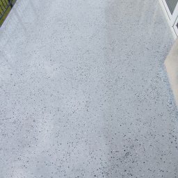 GkRybczyński - Balkon z szarą, błyszczącą posadzką żywiczną z czarnymi i białymi drobinkami, widok z góry, z fragmentem balustrady i okna w tle.