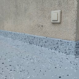 GkRybczyński - Szara, żywiczna posadzka z czarnymi i białymi elementami dekoracyjnymi, wykończona przy ścianie, z widocznym gniazdkiem i zabezpieczeniem folią malarską.